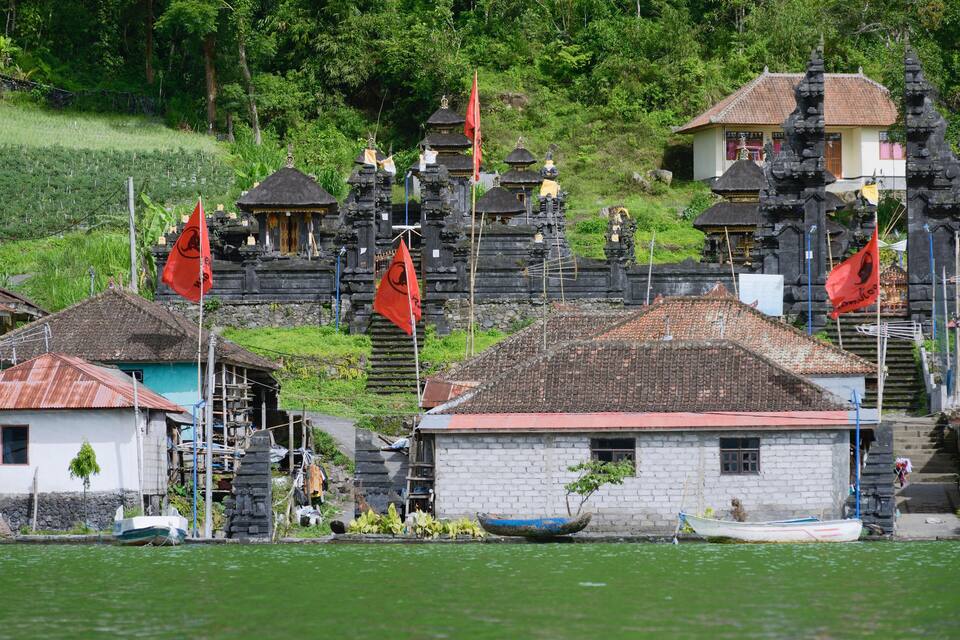 A traditional Balinese temple complex with red flags, a small boat, and a white house by the water.