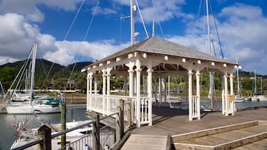 Un pabellón blanco sobre un muelle de madera junto a un puerto deportivo con barcos y una montaña al fondo.