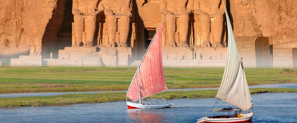 A sailboat on a river with ancient Egyptian temple ruins in the background.
