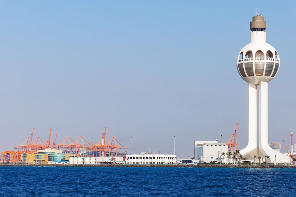 A white tower with a spherical observation deck, a port with orange cranes, and a calm blue sea.