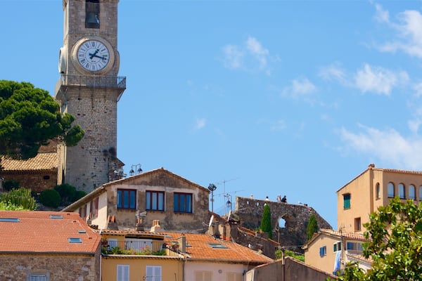 Un beffroi historique, orné de chiffres romains, entouré d’une architecture méditerranéenne typique et surplombé d’un ciel d’un bleu éclatant.
