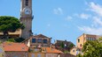 Un beffroi historique, orné de chiffres romains, entouré d’une architecture méditerranéenne typique et surplombé d’un ciel d’un bleu éclatant.