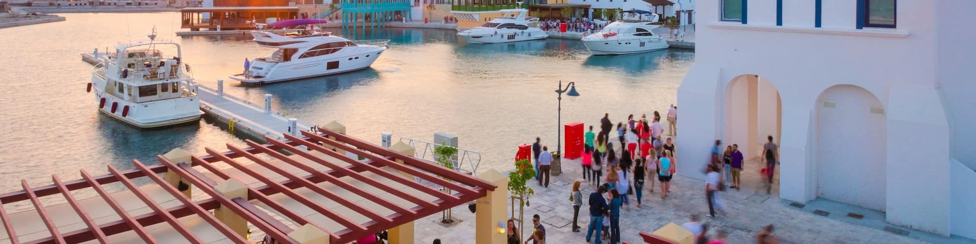 A marina with boats docked, a sunset in the background, and people walking on the promenade.