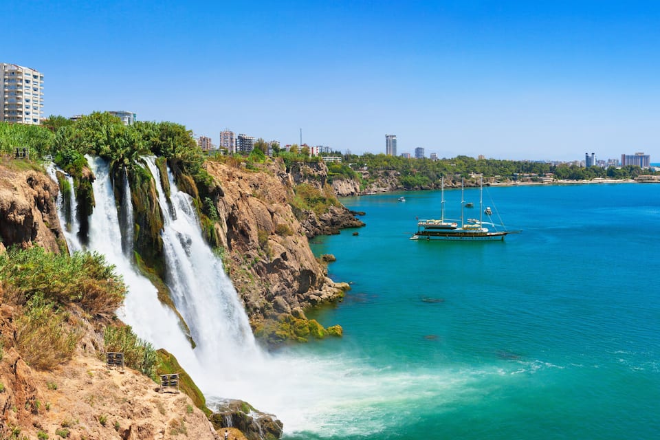 Un paysage urbain côtier avec une cascade imposante, des eaux turquoise et limpides, et un bateau au loin.
