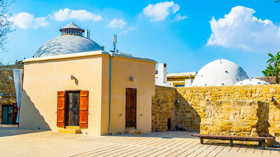 A building with a dome and a smaller structure with a cupola, both with stone walls and wooden shutters, under a clear blue sky.