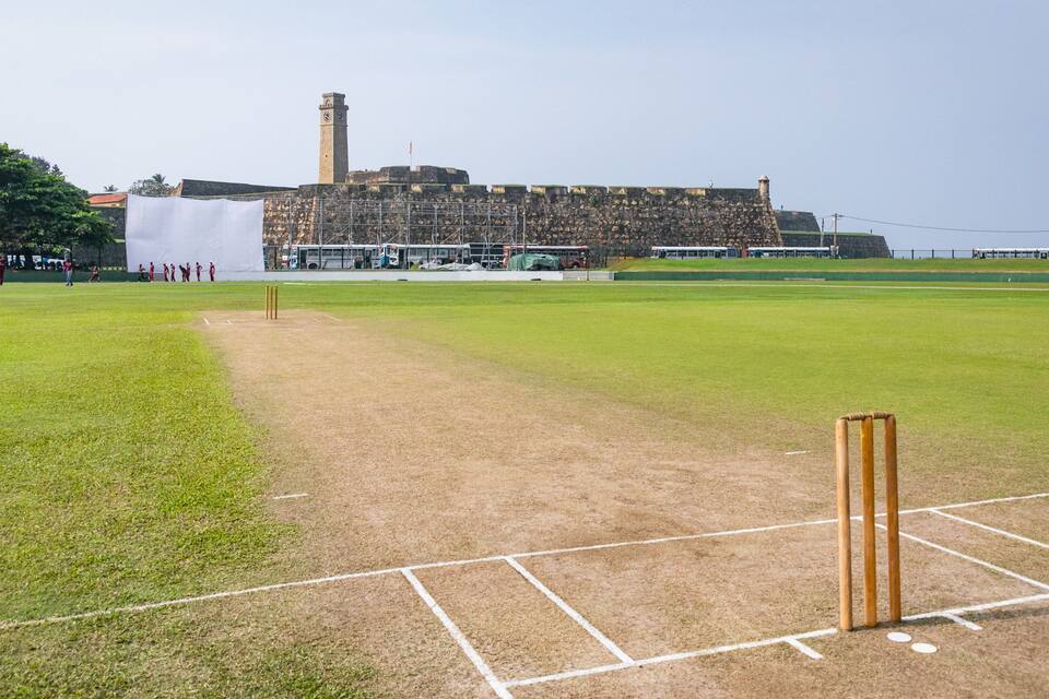 A cricket field with a wicket, a large stone building, and a clock tower in the background.