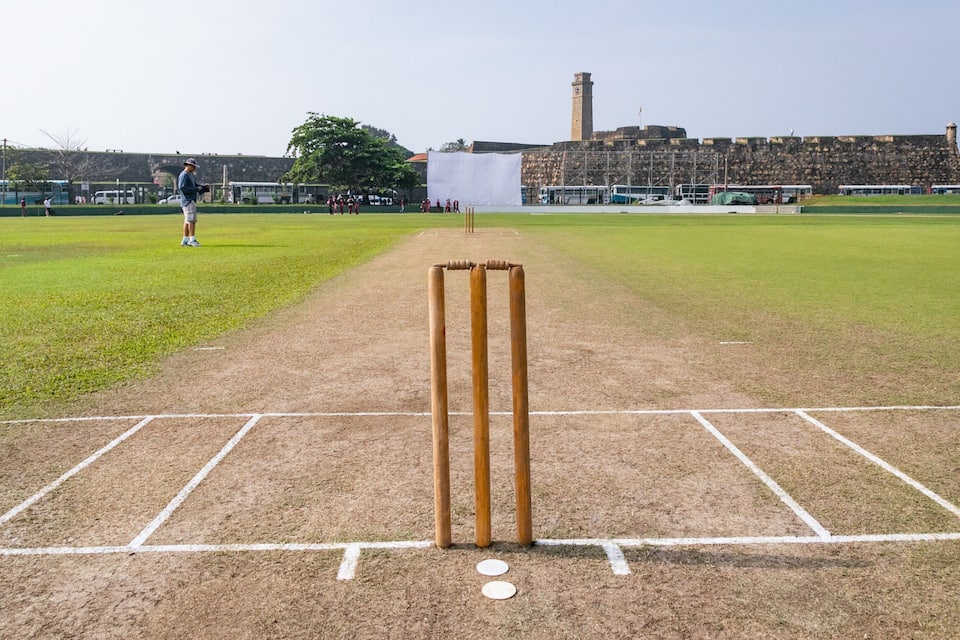 A cricket pitch with stumps in the center, surrounded by a grassy field with a stone wall and a clock tower in the background.