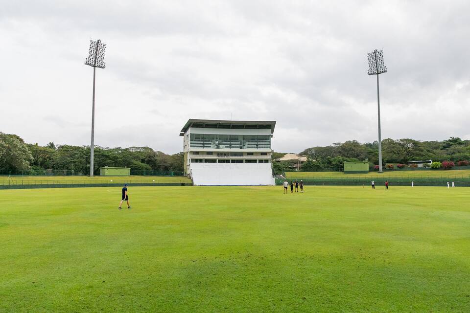 A large, open grassy field with a modern building in the center, surrounded by trees and equipped with tall floodlights.