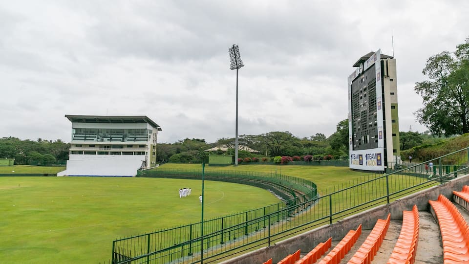 A cricket stadium with green seating, floodlights, and a scoreboard.