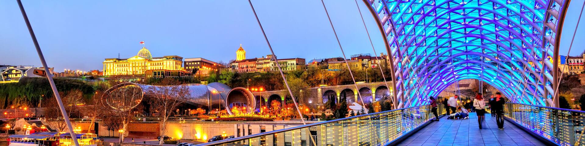 A brightly lit pedestrian bridge with a reflective water surface below, a historic building in the background, and a clear sky.