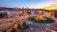 A cityscape at dusk with a roundabout, historic buildings, and a central fountain.