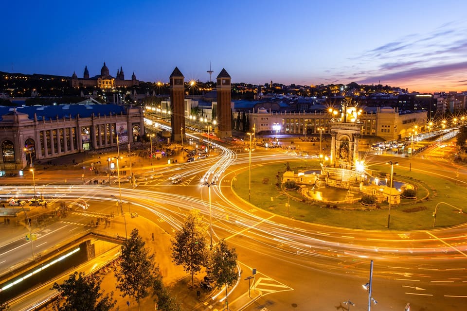 A cityscape at dusk with illuminated buildings, a roundabout, and a prominent monument.