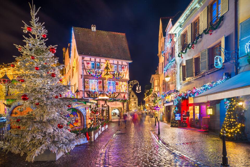Eine Kopfsteinpflasterstraße gesäumt von weihnachtlich dekorierten Gebäuden, darunter ein großer Weihnachtsbaum und beleuchtete Schilder.