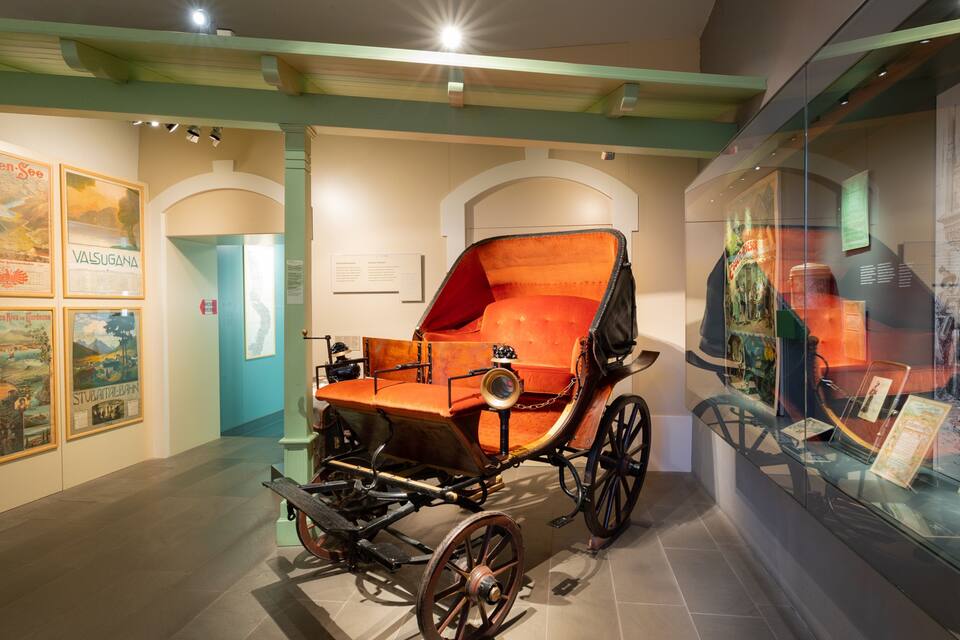 An antique carriage with a red interior, displayed in a museum with historical posters on the walls.