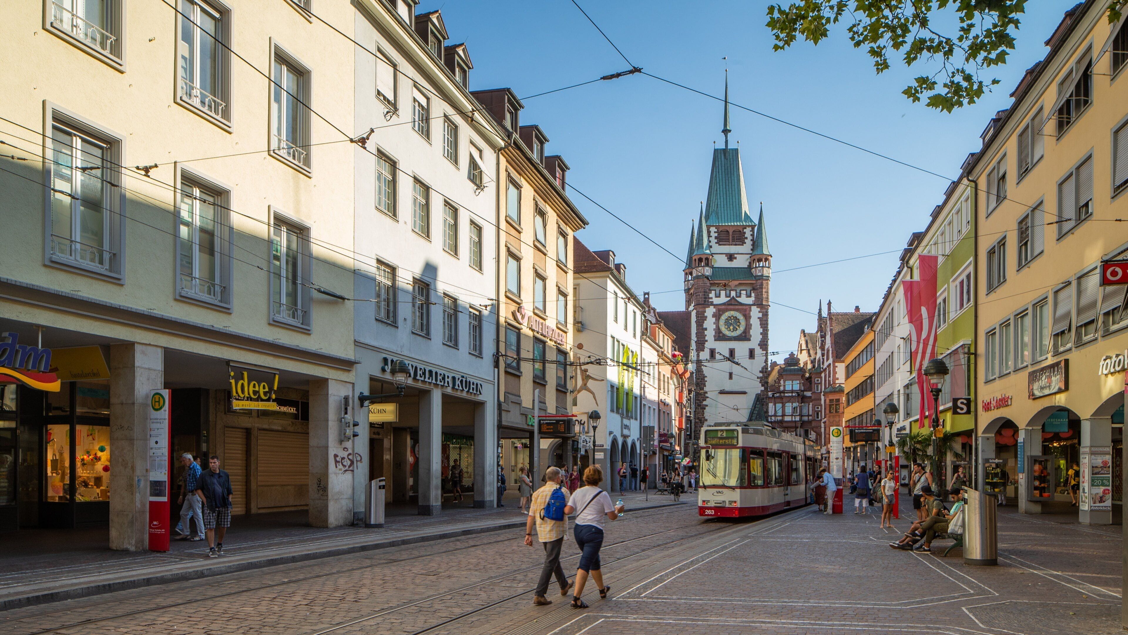 Een tram rijdt over een geplaveid straatje met historische gebouwen en een kerktoren op de achtergrond.