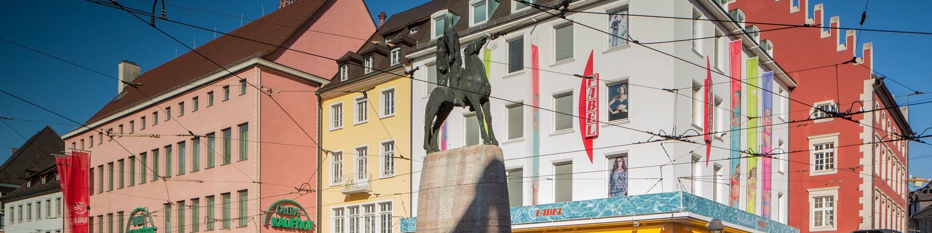 A statue in a square with tram tracks, surrounded by colorful buildings and a tram.