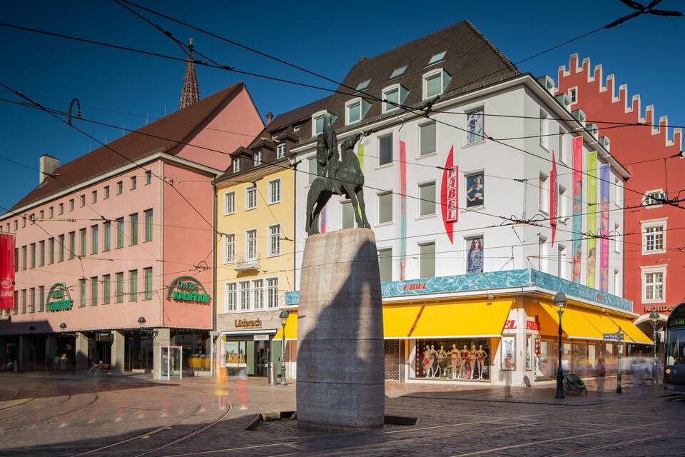 A statue in a square with tram tracks, surrounded by colorful buildings and a tram.