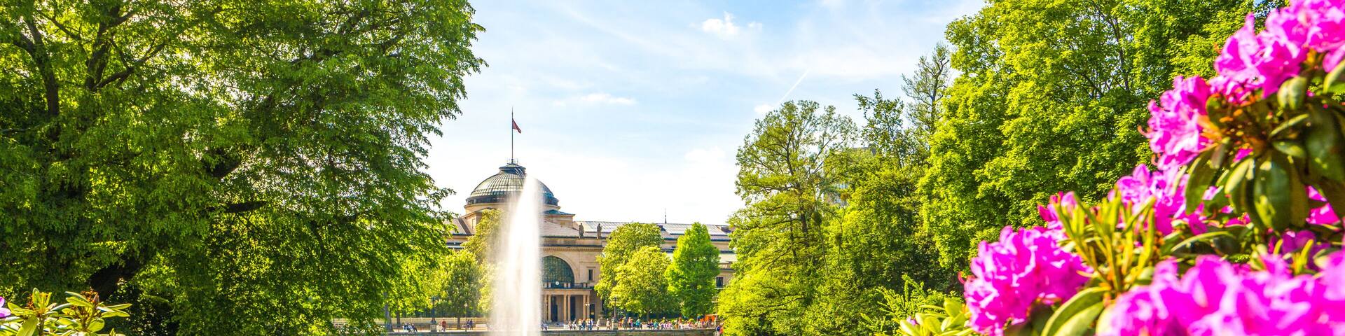 A river with a fountain, surrounded by lush greenery and vibrant pink flowers.