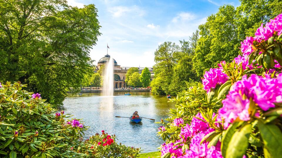 A river with a fountain, surrounded by lush greenery and vibrant pink flowers.