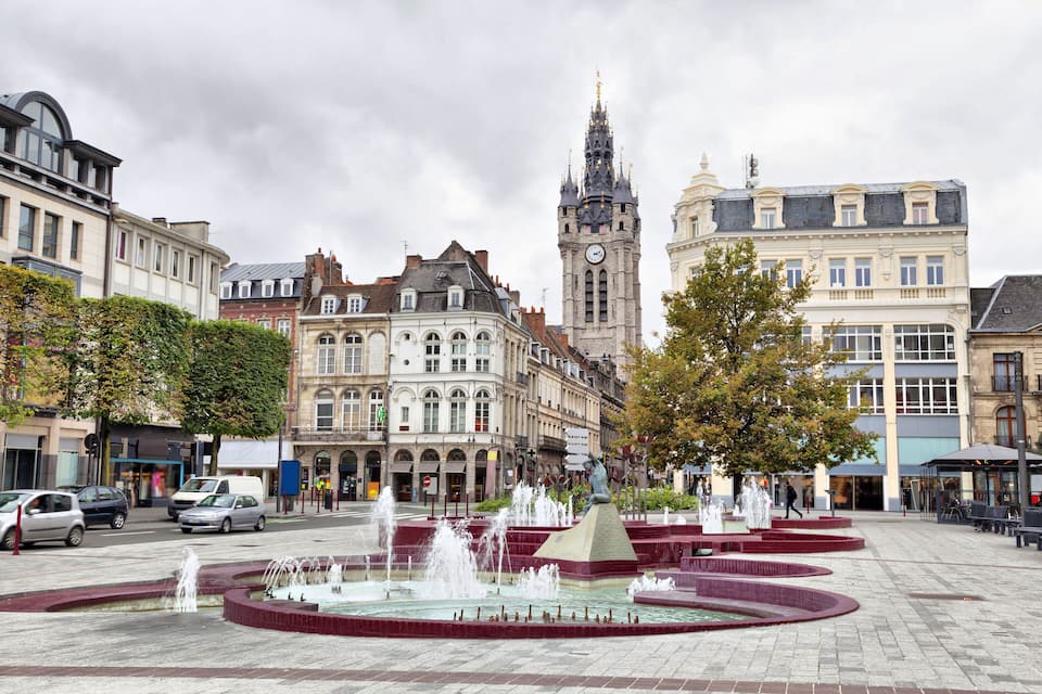 A historic square with a central fountain, a clock tower, and classic European architecture.