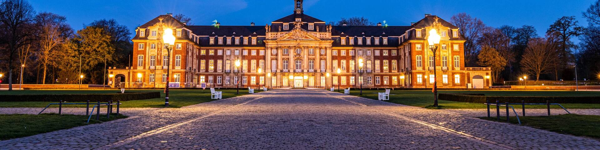 A large, illuminated building with a central dome and symmetrical wings, surrounded by a cobblestone courtyard.
