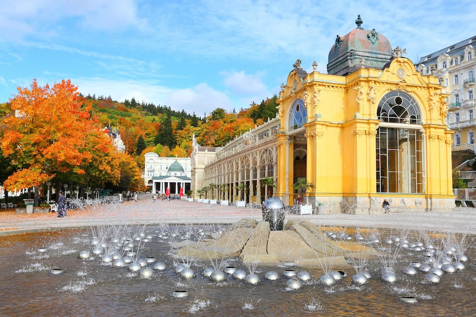A fountain with multiple water jets in a public square, surrounded by autumn-colored trees and historic buildings.
