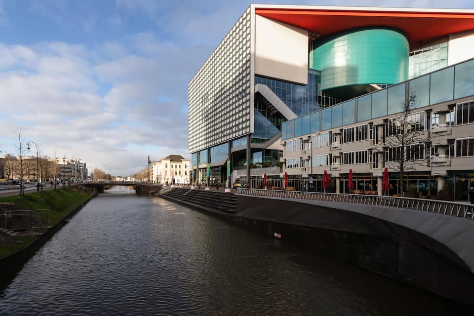 Un edificio moderno con techo rojo y una estructura cilíndrica verde, un río con un puente, y un paisaje urbano al fondo.