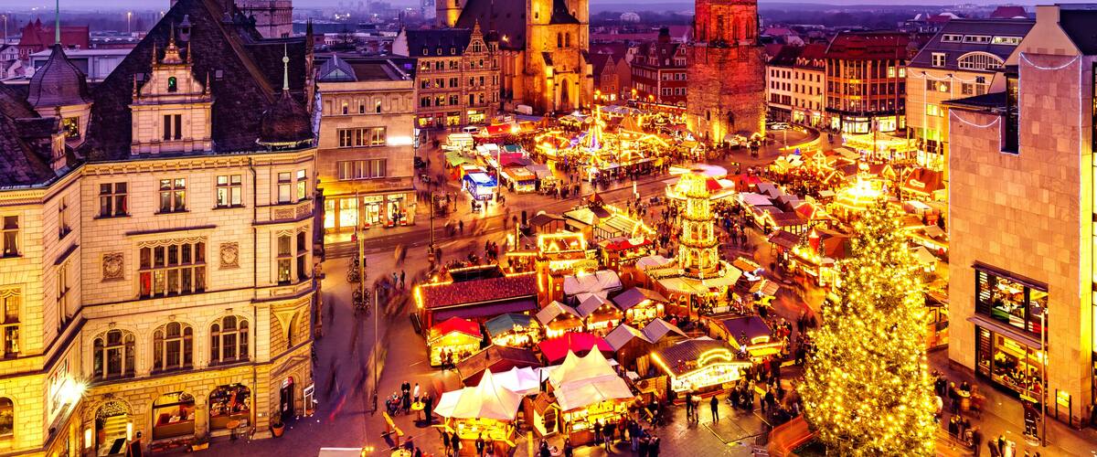 A historic city square with medieval architecture, including a prominent church with twin spires and a clock tower.