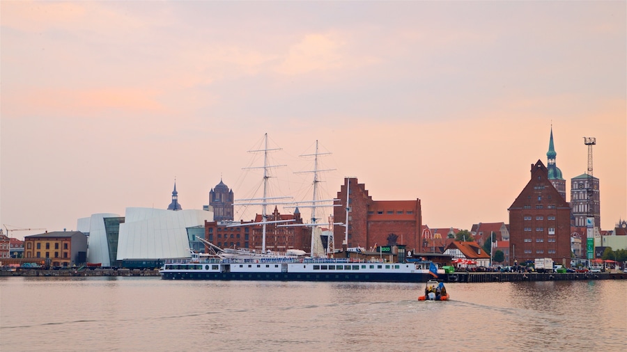 A river with a large cruise ship and a tugboat. In the background, there are historic buildings and a church steeple.