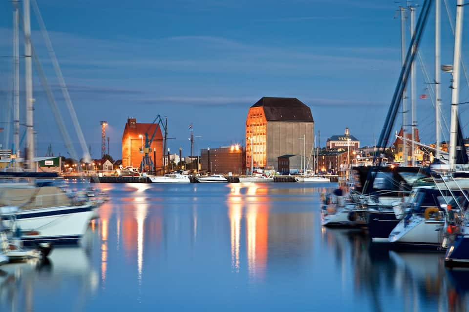 A marina with boats docked, a large building with a sloped roof, and a crane in the background.