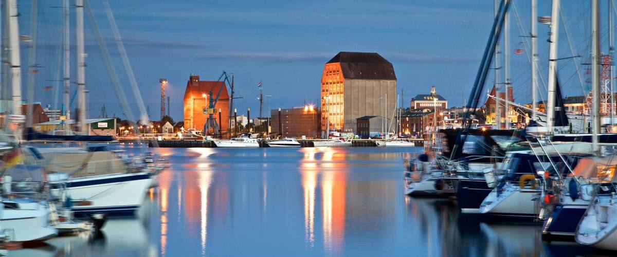 A marina with boats docked, a large building with a sloped roof, and a crane in the background.