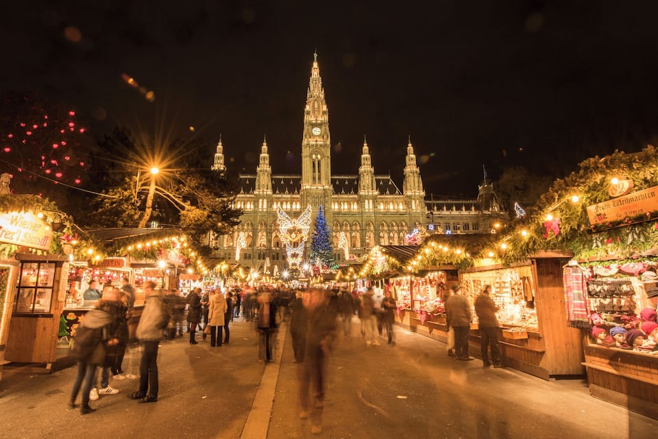 A bustling Christmas market with stalls and people, a prominent church with a tall spire, and festive lights.