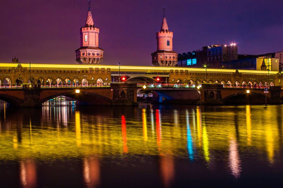 Uma ponte com duas torres e o reflexo de luzes coloridas na água.