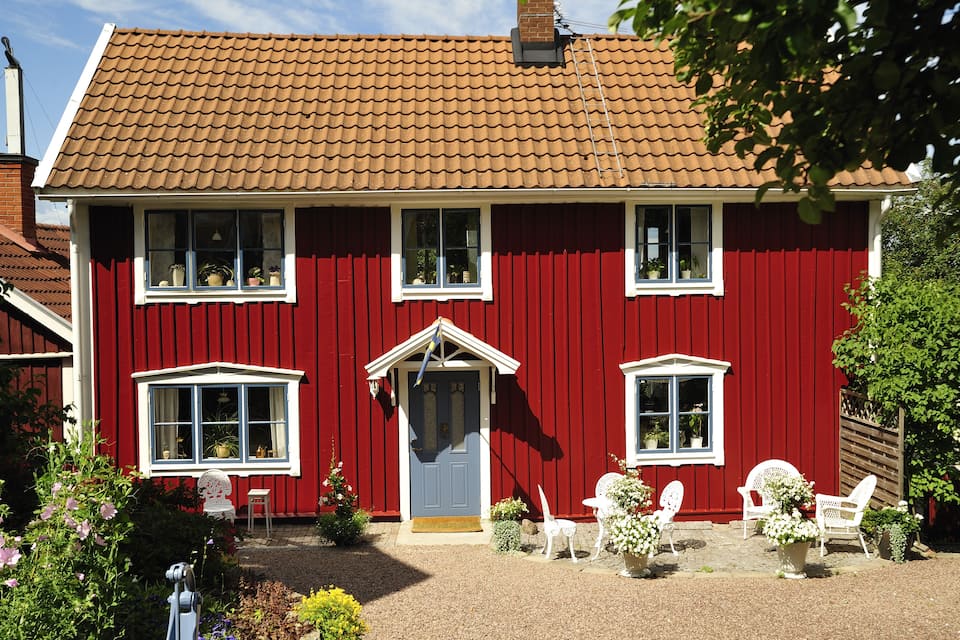 A red house with a brown tiled roof, white trim, and multiple windows. There are white chairs and potted plants in the front yard.