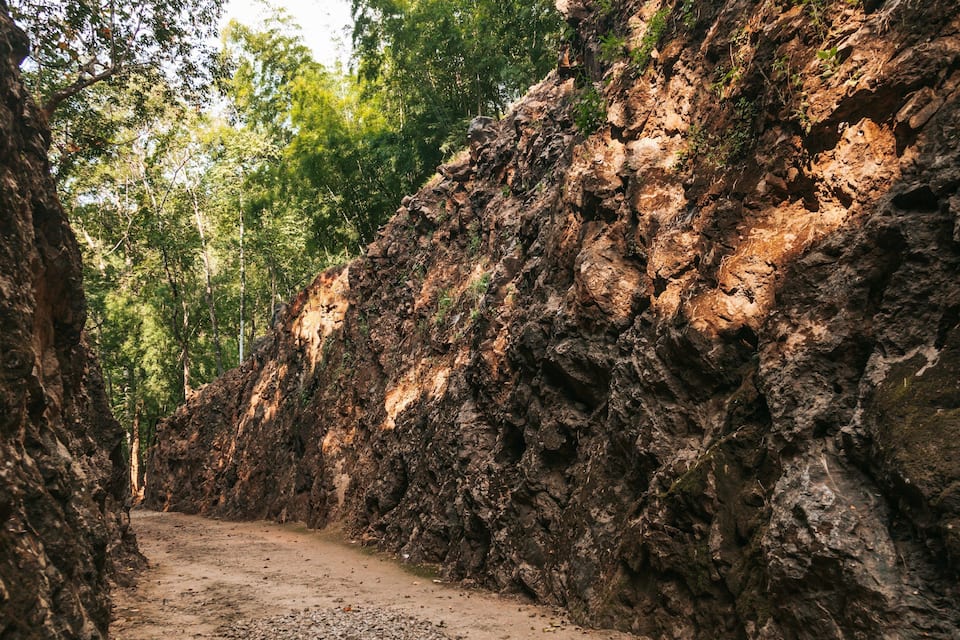 A narrow dirt path flanked by rocky cliffs and dense green foliage.