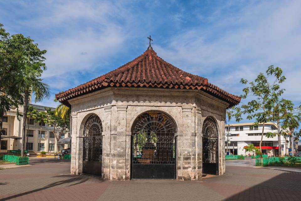 A historic stone gazebo with a tiled roof, surrounded by palm trees and a paved plaza.