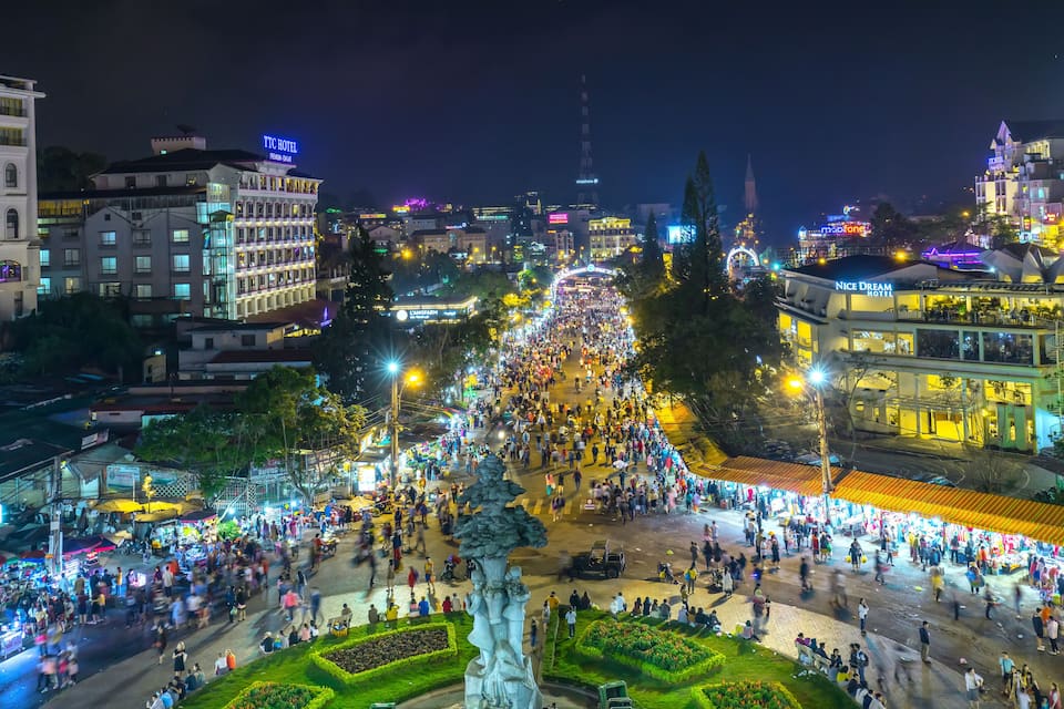 A bustling night market with a large crowd, illuminated buildings, and a prominent statue in the center.