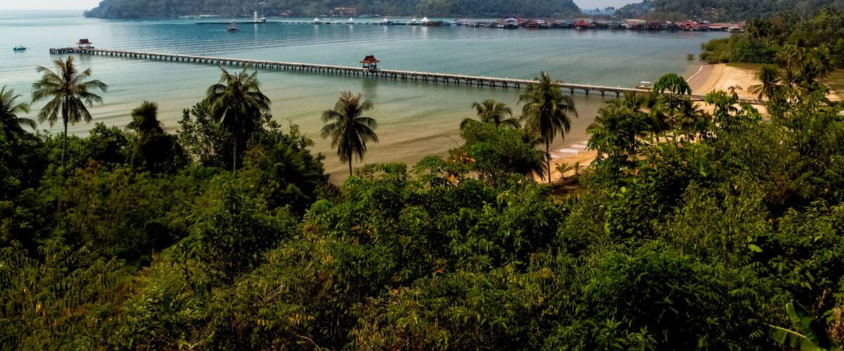 A coastal landscape with a pier extending into the sea, a sandy beach, and lush greenery.
