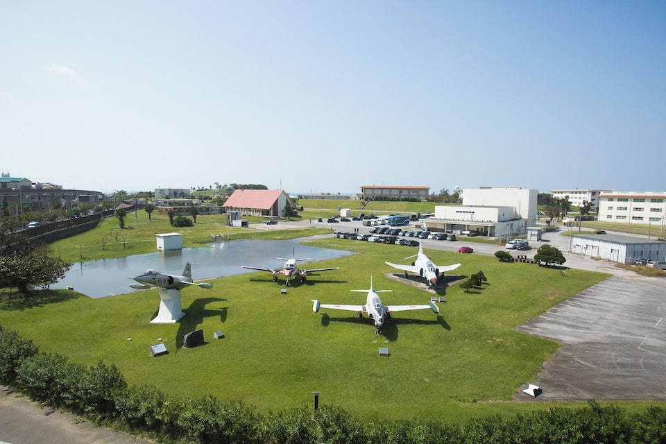 Un camp d'enlairament cobert d'herba amb diversos avions petits aparcats a l'herba, un edifici gran i un pàrquing al fons.