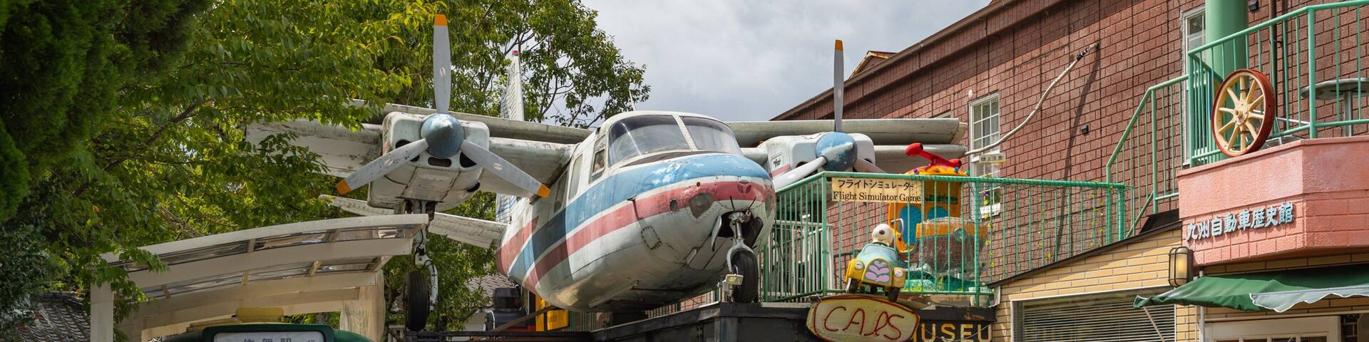 A vintage airplane, a green bus, and a red car in front of a building with a sign that reads "CARS USEU".