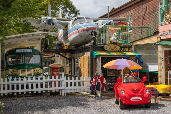 Un avion d’époque, un autobus vert et une voiture rouge devant un bâtiment sur lequel on peut lire l’inscription « CARS USEU ».