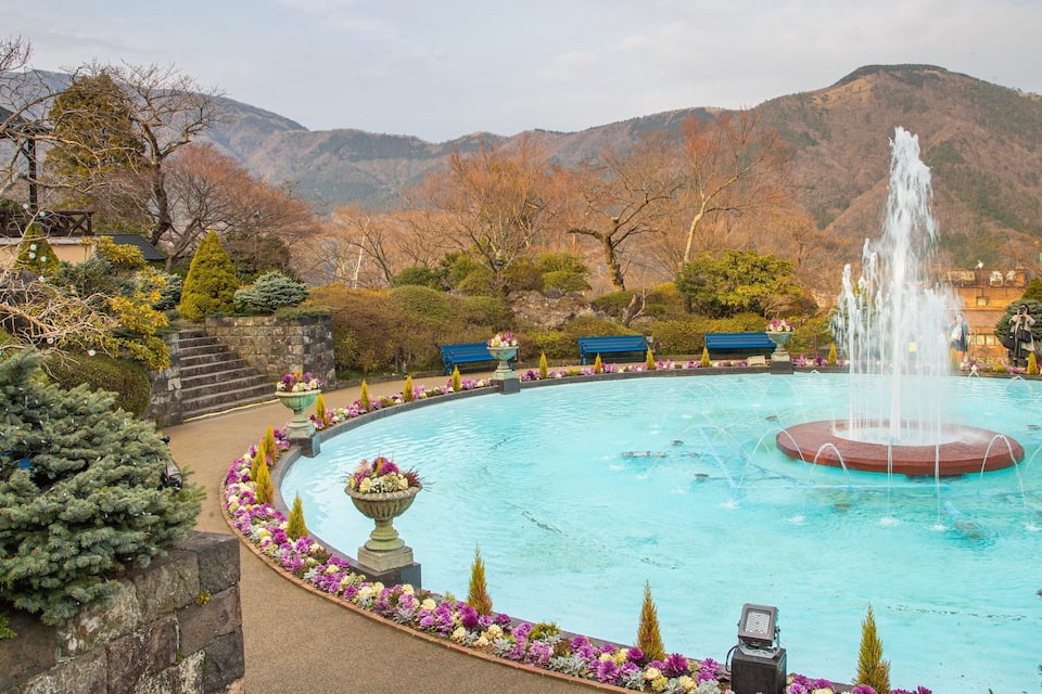 A fountain surrounded by a flower garden, benches, and a mountainous backdrop.