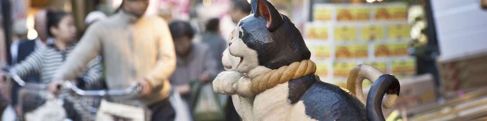 Una escena callejera con una estatua de gato sobre un pedestal de madera, bicicletas y gente al fondo.