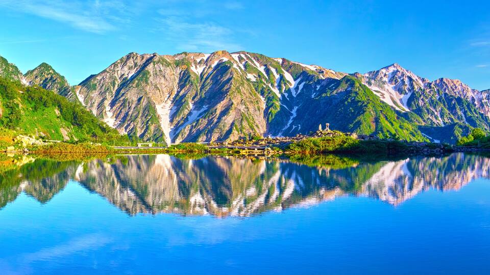 Un lago de montaña con reflejos, colinas verdes y un pequeño edificio.