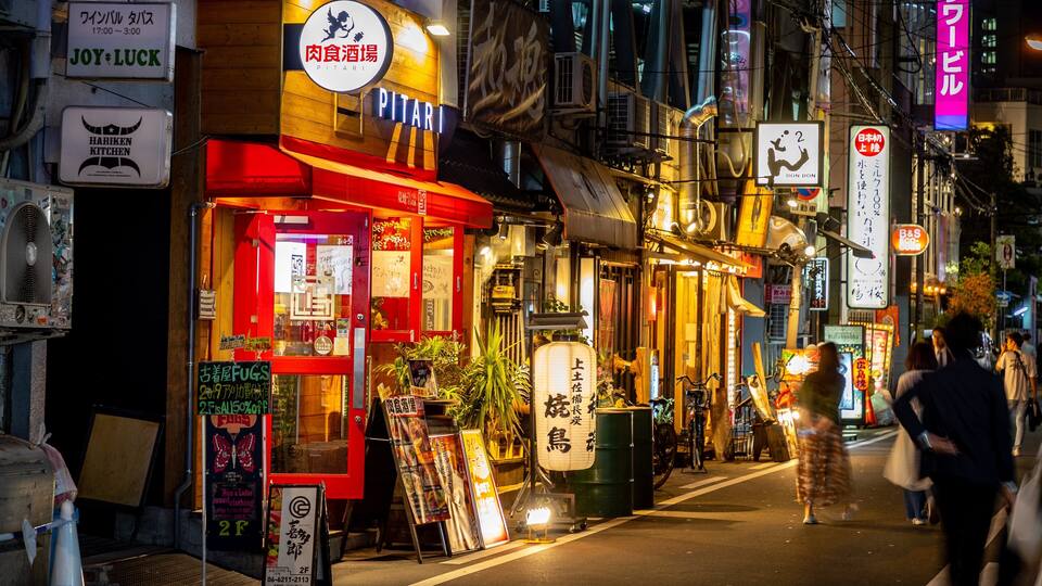 Uma rua movimentada à noite, com diversas lojas e placas, incluindo um restaurante Hariken Kitchen e um restaurante de carnes Pitari.