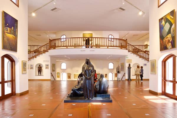 An art gallery with a central sculpture, wooden railings, and various paintings on the walls.