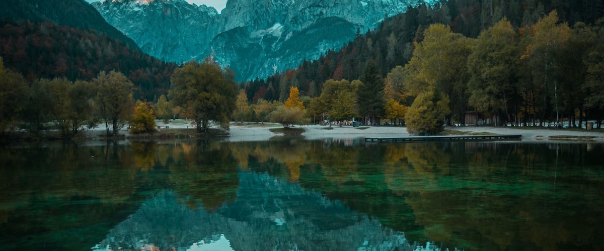 A mountain lake with clear reflections, surrounded by autumn-colored trees and snow-capped peaks.
