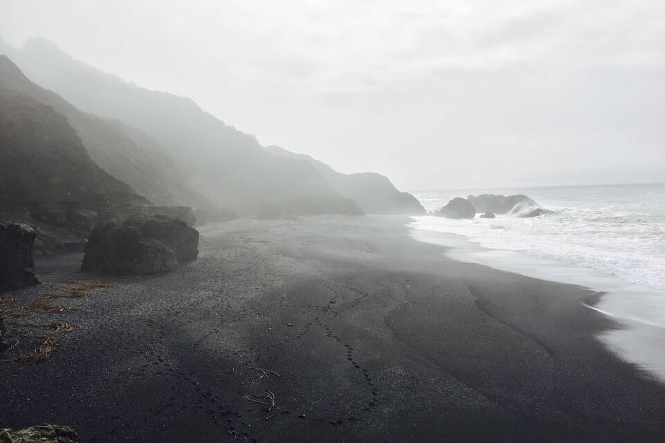 Una playa brumosa de arena oscura y acantilados rocosos.