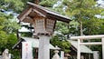 A traditional Japanese torii gate with a stone lantern in front, surrounded by lush greenery and a pagoda in the background.