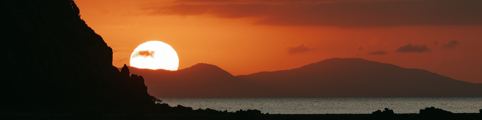 A sunset over a coastal landscape with a prominent cliff and calm sea reflecting the sun.